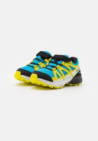 Blue and black athletic shoes with yellow accents. Features textured fabric, Velcro straps, and a rugged rubber sole for traction.