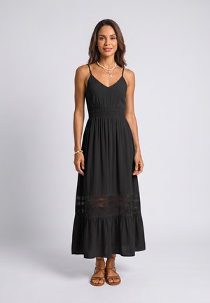 Vrouw draagt een zwarte mouwloze maxi-jurk met kanten detail, bruine sandalen en gelaagde parelkettingen, staand tegen een effen lichte achtergrond.