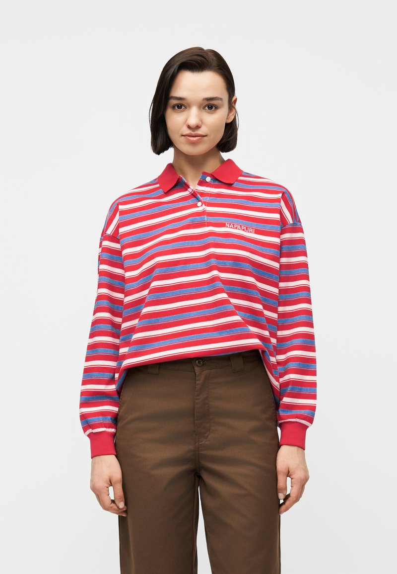 Γυναίκα που φοράει μακρυμάνικη πόλο μπλούζα με κόκκινες, λευκές και μπλε ρίγες και καφέ παντελόνι, στέκεται μπροστά σε ένα απλό φωτεινό φόντο.