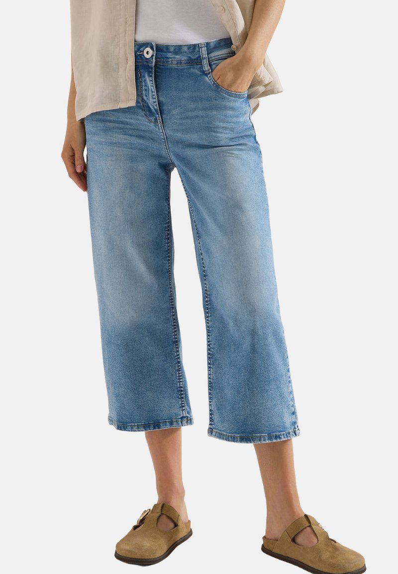 Personne portant un jean large raccourci bleu clair et des chaussures beige à enfiler, une main dans la poche et une chemise beige par-dessus un top blanc.