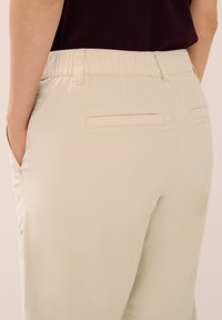 Světlé béžové kalhoty s elastickým pasem, dvěma zadními kapsami a hladkou texturou tkaniny. Obsahují jemné detaily šití.
