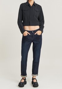 Lyhyempi musta napillinen takki, jossa on rintataskut, yhdistetty tummansinisiin kapeneviin farkkuihin ja mustiin platform-korkokenkiin, joissa on nilkkahihnat.