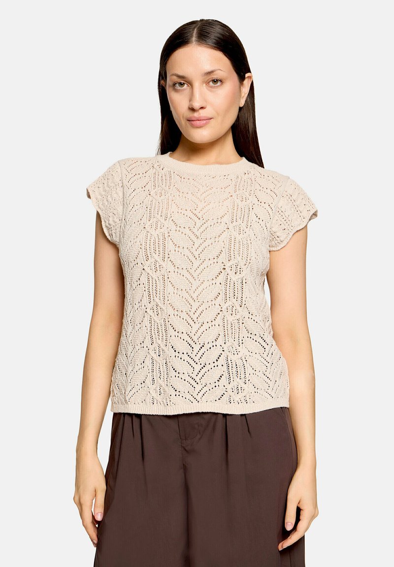 Femme aux cheveux raides foncés portant un haut en maille beige à manches courtes avec un motif en dentelle et un pantalon ample marron foncé sur fond blanc.