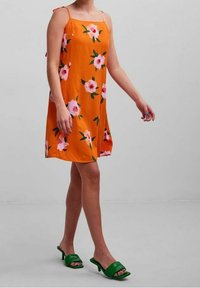 Orange floral dress with thin straps, featuring pink and green flowers. Model wears green slide sandals, showcasing a relaxed fit and flowy design.