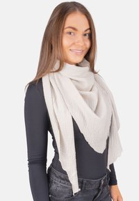 Jeune femme souriante, portant une grande écharpe texturée beige clair sur un haut noir à manches longues ajusté et un jean foncé, posant devant un fond blanc.