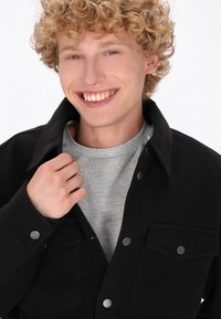 Giacca nera con due tasche frontali e bottoni a pressione argentati, indossata sopra una camicia grigia chiaro. Il modello ha capelli biondi ricci e sta sorridendo.