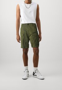 Olivgrön cargo shorts med flera fickor, en framdragbar dragkedja och en avslappnad passform, tillsammans med en vit ärmlös skjorta och svarta och vita sneakers.