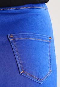 Blå denimjeans med en bakficka med orange sömmar, slät textur och en figurnära design som framhäver plaggets form.