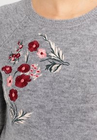 Pull gris tricoté avec des fleurs rouges et roses brodées et des feuilles vertes près de l'encolure.