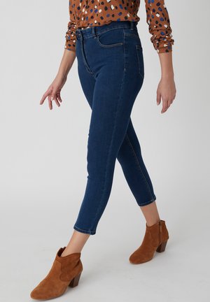 Femme portant un jean court bleu marine, des bottines en suède marron et une chemise à manches longues marron à motif, marchant sur un fond uni.