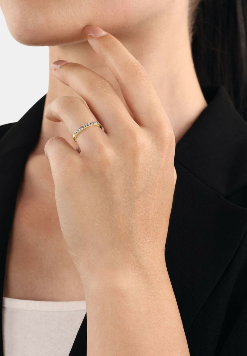 Anello d'oro con una fila di pietre preziose trasparenti lungo la parte superiore, indossato su una mano che tocca il mento, su uno sfondo neutro.