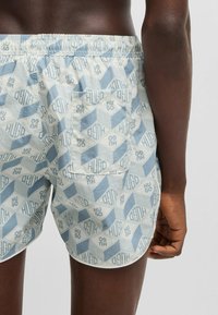 Pantalones cortos de baño en azul claro y crema con patrones geométricos. Cintura elástica, bolsillo trasero y una tela ligera y texturizada.