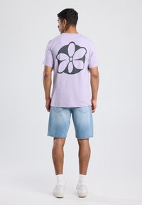 Lila t-paita, jossa on suuri musta kukkakuva selässä, yhdistettynä vaaleansinisiin farkkushortseihin ja valkoisiin lenkkareihin. Yksinkertainen ja rento asu.