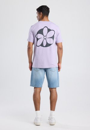 Lila t-shirt met een grote zwarte bloemenafbeelding op de achterkant, gecombineerd met lichtblauwe spijkershorts en witte sneakers. Eenvoudige en casual outfit.