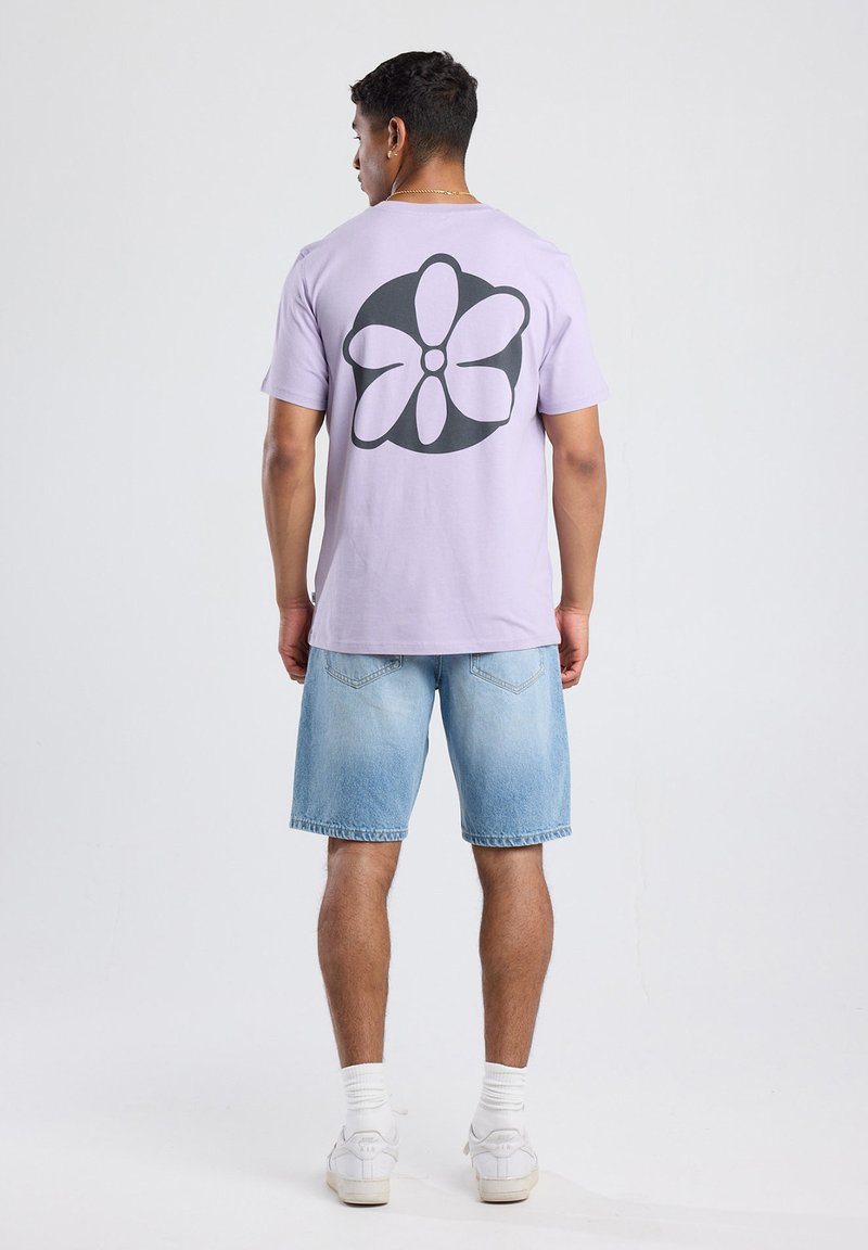 Lila t-paita, jossa on suuri musta kukkakuva selässä, yhdistettynä vaaleansinisiin farkkushortseihin ja valkoisiin lenkkareihin. Yksinkertainen ja rento asu.