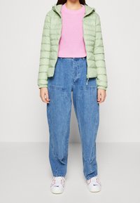 Hellgrüne gepolsterte Jacke mit Kapuze, pinker Rippstrickpullover, lockere blaue Jeanshose und weiße Sneakers mit roten Akzenten.