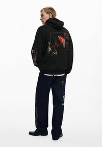 Sort hættetrøje med livlige, broderede designs på ryggen og ærmerne; parret med mørkeblå jeans med patchwork-detaljer.