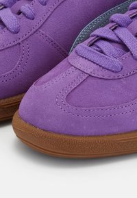 Gros plan sur des baskets en daim violet avec des semelles en caoutchouc marron texturées et des lacets violets, montrant des coutures détaillées et une partie d'un accent gris.