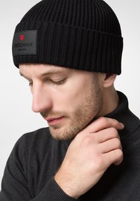 Czarna dzianinowa czapka z prążkowanym splotem oraz prostokątną metką z czerwoną logo. Noszona przez osobę w ciemnym ubraniu, widok z bliska.