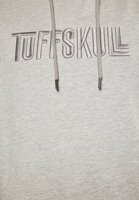 Szürke kapucnis pulóver, kiemelkedő "TUFFSKULL" felirattal fémes ezüst színben. Kábelhúzó fém végződésekkel. Puha pamut keverékből készült.