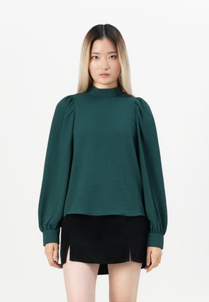 Femme avec de longs cheveux blonds raides portant un haut vert foncé à manches volantes et une mini jupe noire avec des fentes sur le côté, se tenant devant un fond blanc.