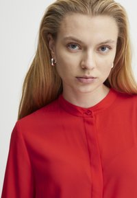 Blusa rossa a maniche lunghe con colletto e chiusura frontale con bottoni, realizzata in un tessuto liscio. Il modello indossa orecchini a cerchio in argento.
