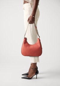 Borsa a tracolla rossa in pelle con un design arrotondato, superficie texture e tracolla grigia. Il modello indossa pantaloni bianchi e décolleté neri a punta.