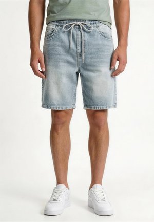 Muž v svetlomodrých riflových šortkách s sťahovacou šnúrkou v páse a bielych teniskách, stojaci na jednoduchom bielom pozadí.