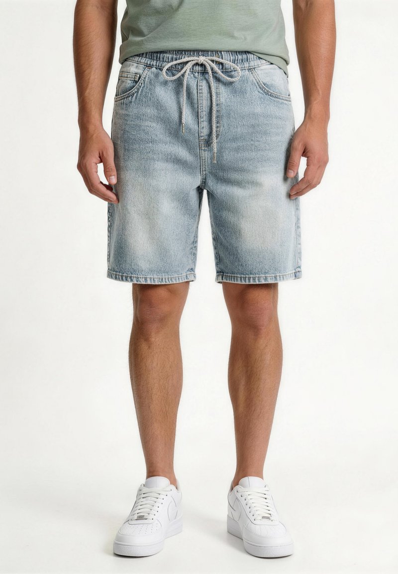 Άνδρας που φοράει ανοιχτό μπλε τζιν σορτς με κορδόνι στη μέση και λευκά αθλητικά παπούτσια, στέκεται μπροστά σε απλό λευκό φόντο.