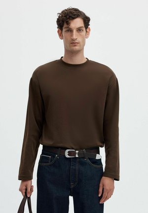 Homme aux cheveux bouclés et foncés portant une chemise marron à manches longues et un jean foncé, tenant une sangle de sac marron devant un fond uni.