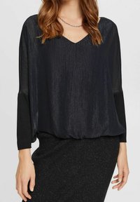 Blusa nera con scollatura a V, maniche lunghe trasparenti e un tessuto testurizzato che crea un effetto scintillante. Abbinata a una gonna scura e luccicante.