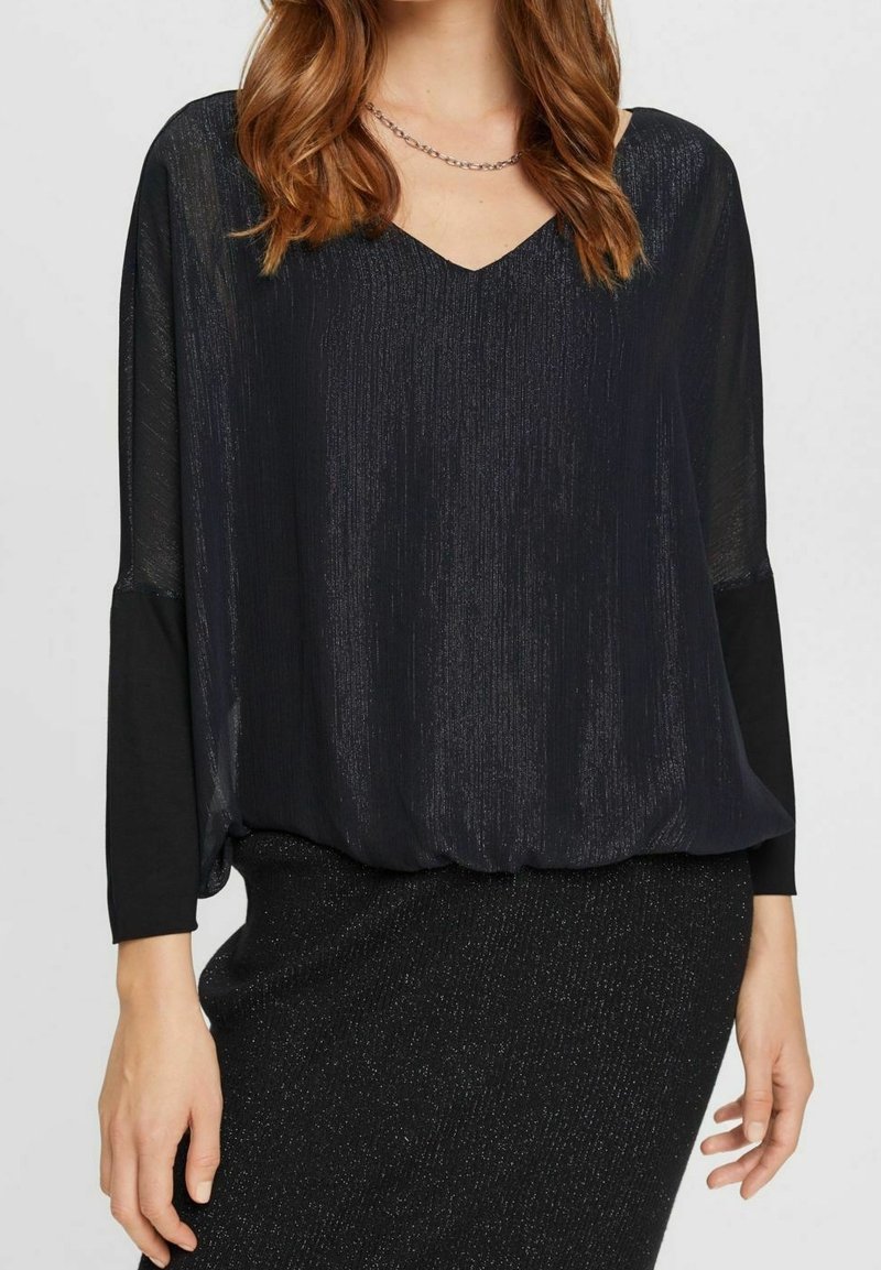 Blusa nera con scollatura a V, maniche lunghe trasparenti e un tessuto testurizzato che crea un effetto scintillante. Abbinata a una gonna scura e luccicante.