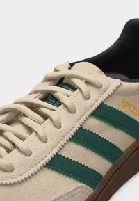 Μπέζ σουέντ αθλητικό παπούτσι με σκούρες πράσινες ραβδώσεις με υφή, κρέμ κορδόνια, μαύρη εσωτερική επένδυση και το "SPEZIAL" σε κίτρινο στο πλάι.