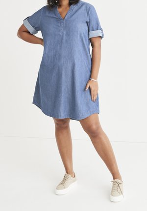 Kobieta w niebieskiej jeansowej sukience z podwiniętymi rękawami stoi z jedną ręką na biodrze i nosi beżowe trampki na platformie na białym tle.