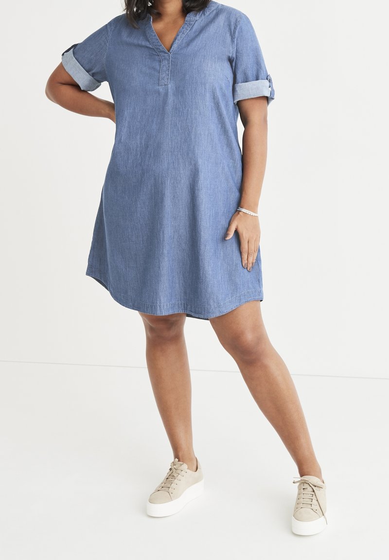 Žena v modrých denimových šatech s ohrnutými rukávy stojí s jednou rukou na boku a má na sobě béžové platformové tenisky na bílém pozadí.