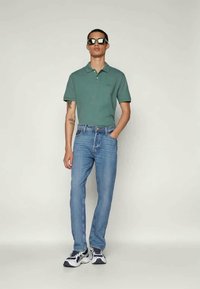 Camisa polo verde, jeans azul claro e ténis brancos. O modelo está posicionado contra um fundo claro, com óculos de sol que adicionam um detalhe elegante.