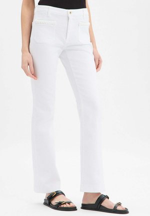 Frau trägt weiße, gerade geschnittene Hosen mit aufgesetzten Taschen vorne und schwarze Pantoletten mit goldenen Schnallen.