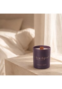 Bougie parfumée à la rose en velours violet avec une mèche en bois sur une table de chevet en pierre beige près du lit et un rideau transparent.