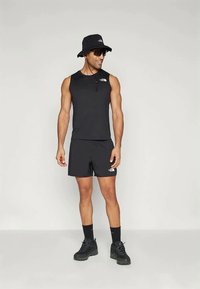 Άνδρας που φοράει μαύρο αμάνικο τοπ, σορτσάκια, καπέλο μπουκάλα, γυαλιά ηλίου, μαύρες κάλτσες και ορειβατικά παπούτσια, στέκεται μπροστά σε απλό ανοιχτό φόντο.