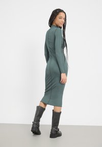 Green ribbed long-sleeve dress with fitted silhouette and scalloped hem. Paired with knee-high black leather boots featuring metallic accents.
