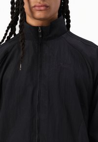 Chaqueta de nylon negra con cuello alto y cierre de cremallera; presenta un sutil patrón texturizado y un detalle del logo en el pecho.