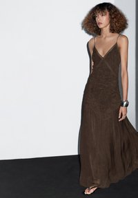 Woman with curly hair wearing a long brown lace-trimmed dress and metallic cuff bracelet, standing against a plain white and black background.