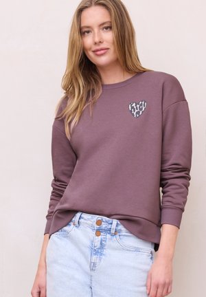 Jeune femme aux longs cheveux blonds portant un sweat mauve avec un petit cœur à imprimé léopard et un jean taille haute bleu clair.
