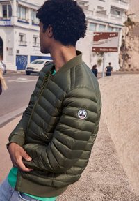 Uomo in giacca trapuntata verde con logo sulla manica si appoggia a un muro di pietra in una strada urbana, con persone e edifici sfocati sullo sfondo.