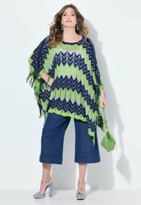 Färgglad poncho i marinblått och limegrönt, med ett zigzag-mönster och fransdetaljer. Tillsammans med vida jeanstights och silverfärgade klackar.