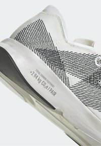 Zapatilla deportiva blanca con patrones lineales negros, tejido texturizado y suela gris. Presenta una etiqueta de huella de carbono cerca del talón.