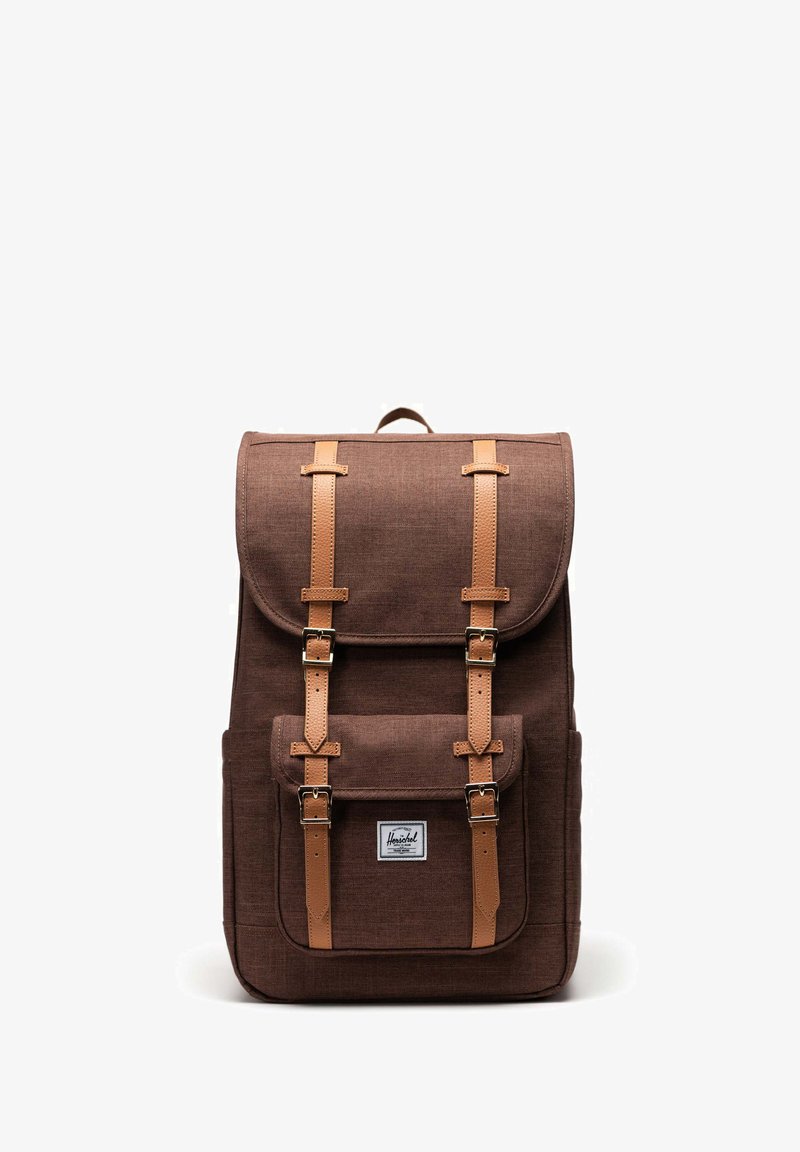 Hnědý látkový batoh se strukturovaným tvarem, vybavený koženými popruhy, kovovými sponami, přední kapsou a víkem nahoře.