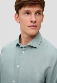 Jasnozielona lniana koszula z wyciętym kołnierzykiem, z zapinanymi na guziki przodem i delikatną fakturą. Widoczne głowa i ramiona modela.