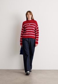 Rood en roze gestreepte gebreide trui met een geribbelde zoom, gecombineerd met donkere spijkerbroek en zwarte schoenen, met een blauwe handtas.