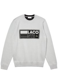 Jasnoszary sweter z okrągłym dekoltem z czarno-białym prostokątnym logo Lacoste oraz zielonym emblematem krokodyla umieszczonym na środku klatki piersiowej.
